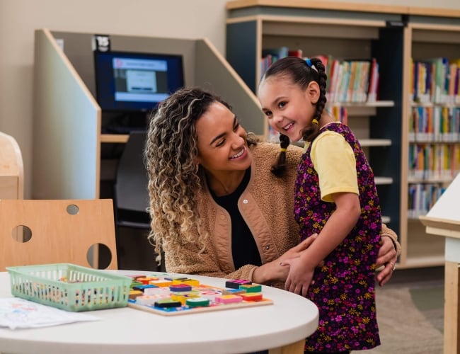 A mother and daughter standing by a table and play with a puzzle in a ibrary