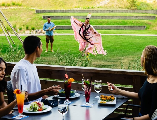 Three individuals sitting at an outdoor table while an Indigenous dancing demonstration takes place in the background