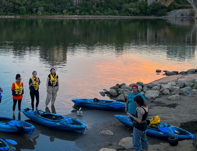 6 people standing along the river with kayaks and life jackets