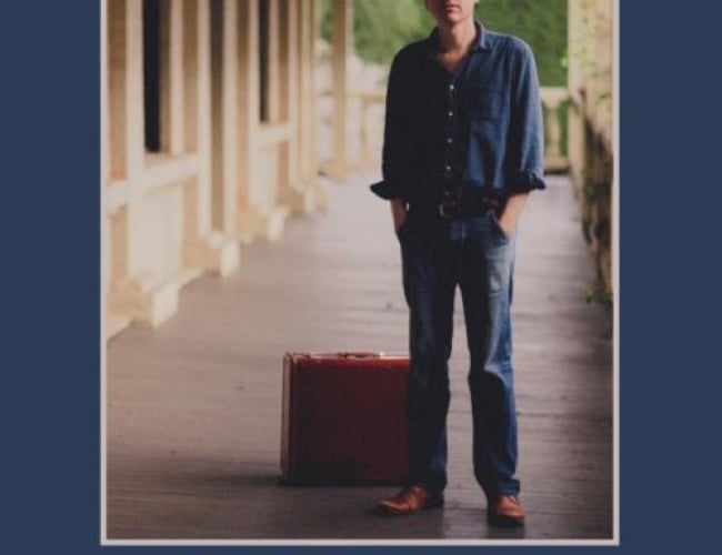 Image of Joel Plaskett standing with briefcase on the floor behind him. Background is unclear of where but it is a building with a sort of porch allowing for a view of a hedge in the back. Text on poster says: "JOEL PLASKETT, ONE REAL REVEAL ON WHEELS. October 3,2024. The broadway Theatre. Tickets at broadwaytheatre.ca. At the very bottom of the poster is a broadway theatre logo and a QR code to buy tickets. 