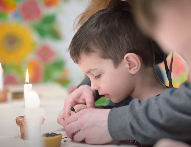 an adult helps a young child write with beeswax on a Ukrainian Easter egg (pysanka)