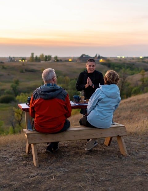 Couple sitting at an outdoor patio table set with food and drinks, while a host speaks to them