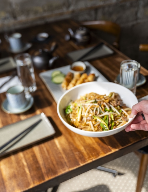 A person holding a bowl of noodles over a table.