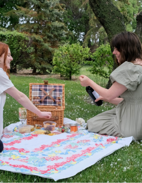 Two people sitting on a blanket, while one opens a bottle of wine.